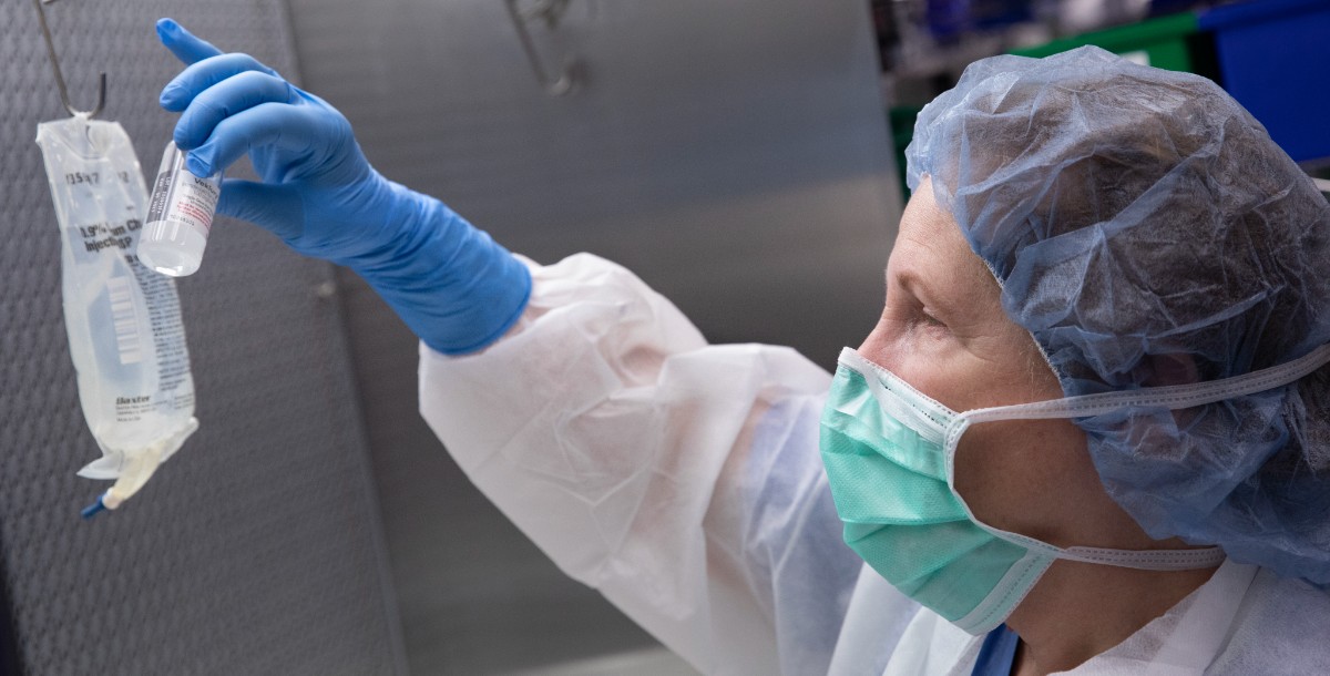 A masked, female pharmacy technician wearing various items of personal protective equipment prepares remdesivir for a patient in the pharmacy.