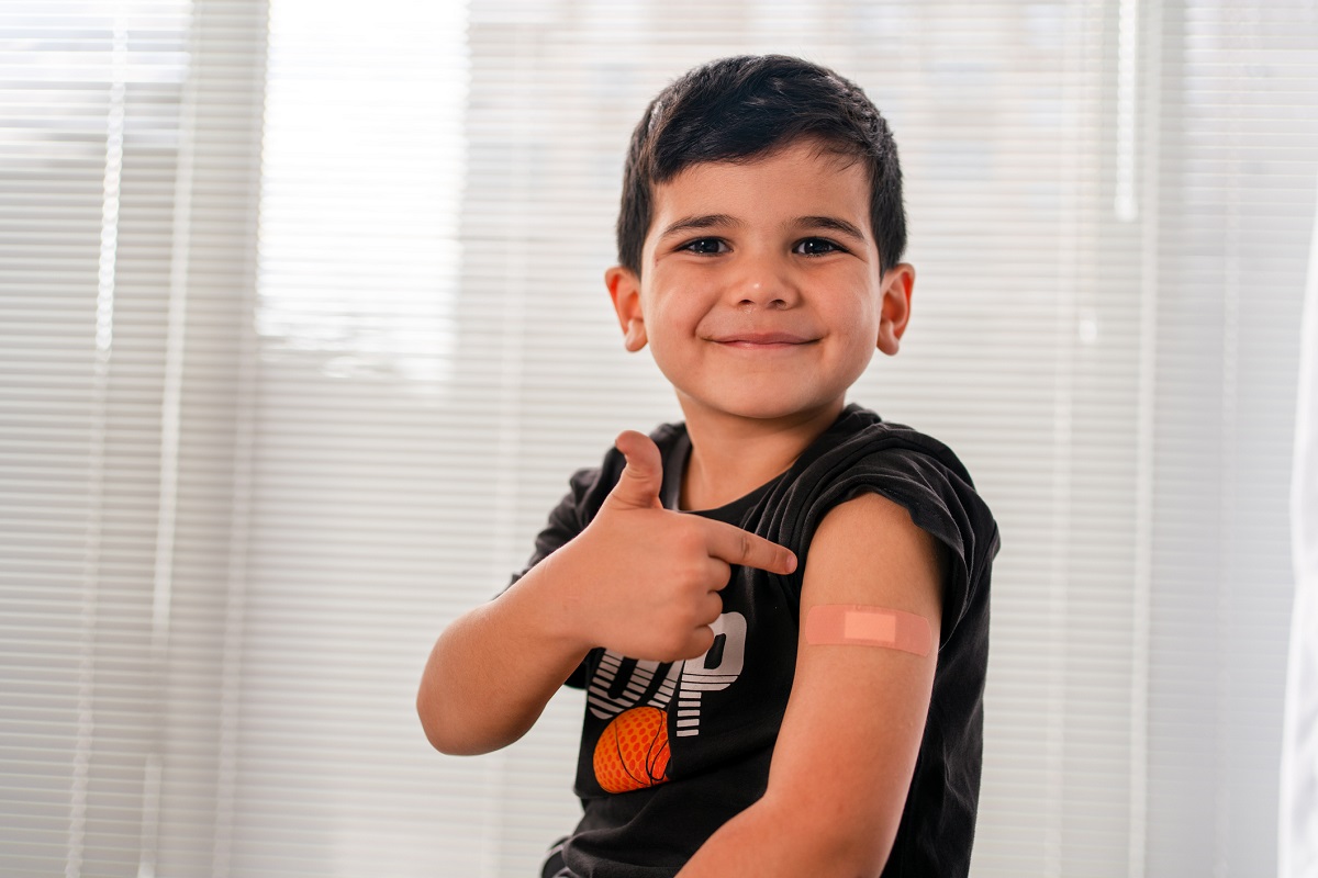 A vaccinated child points to an adhesive bandage on his arm, with his shirt sleeve rolled up.