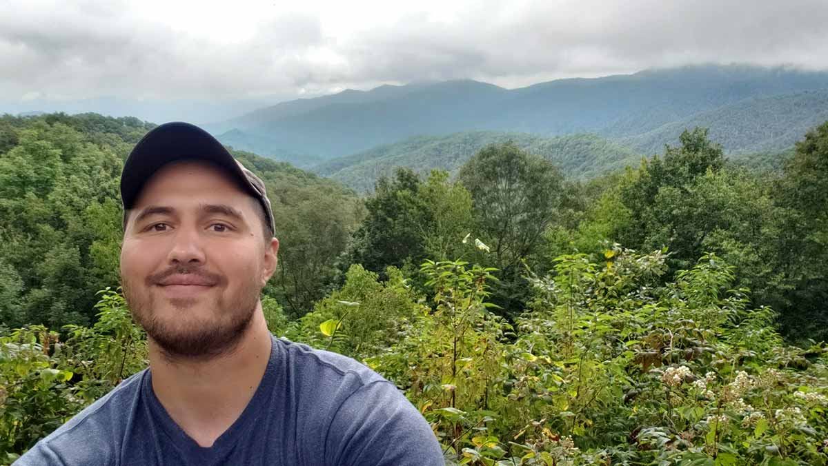 A young person is standing outdoors in front of foliage with a view of mountains and clouds in the distance.