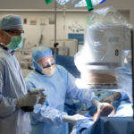 Two doctors in personal protective equipment stand next to an operating table with a patient on it.