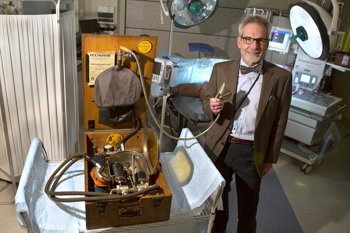 Man smiling, standing in an operating room, holding a face mask from an antique piece of anesthesiology equipment.