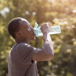 An overheated man drinks water from a bottle in a park.