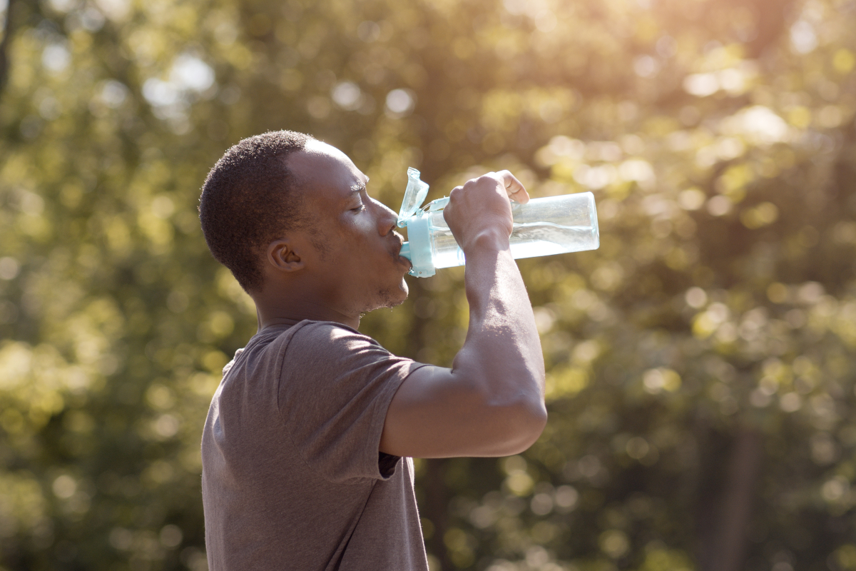 An overheated man drinks water from a bottle in a park.