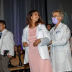 An incoming medical student smiles as her white coat is put on, with a faculty member behind her.