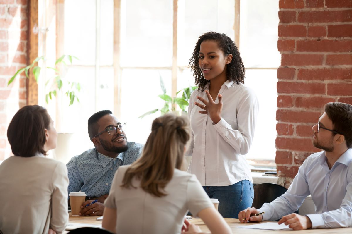 Employees of various ethnicities sitting around an office table, listening to one employee who is standing and speaking