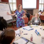 Women sit around round table during a conference while a woman stands at a large notepad.