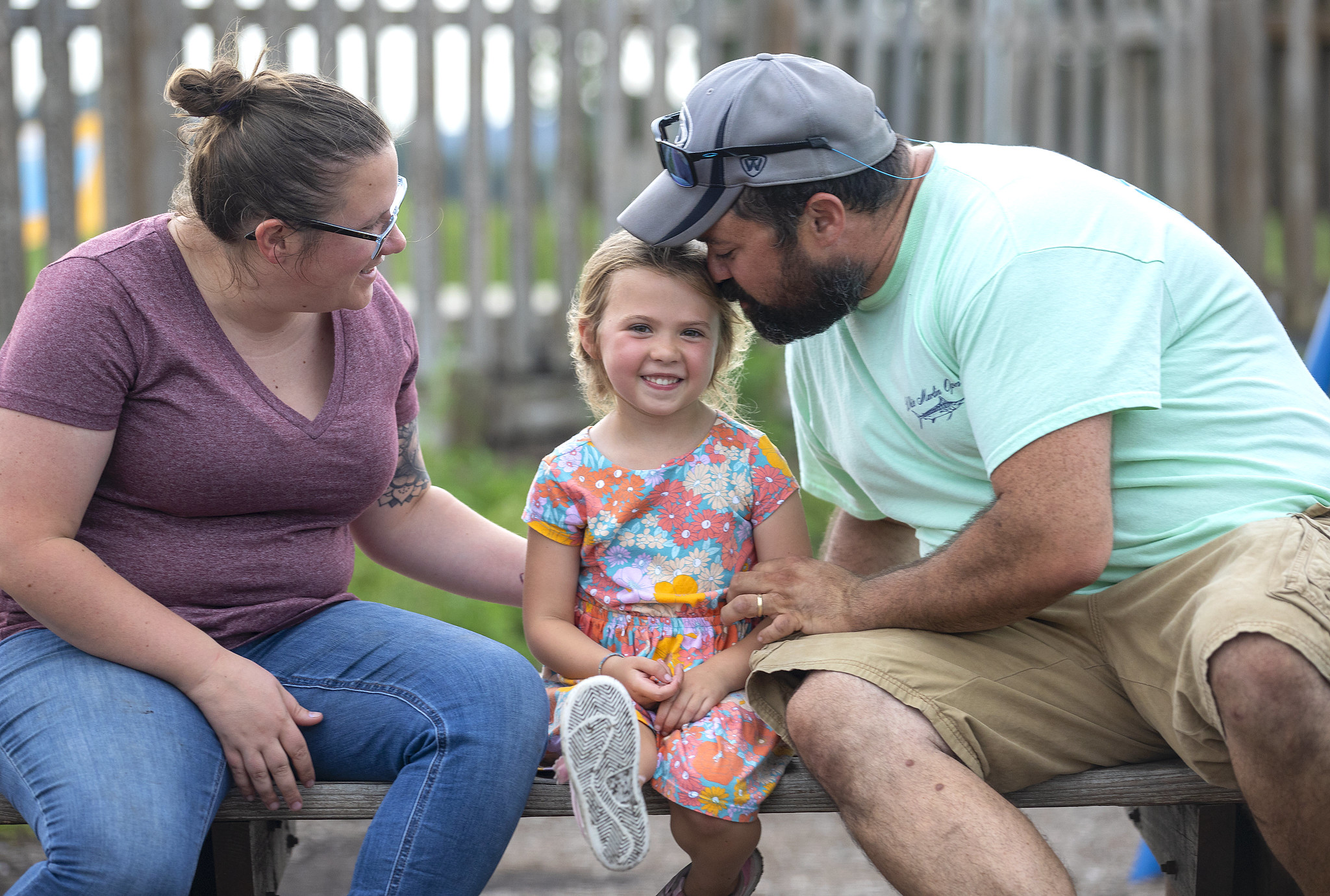 A man and a woman to the left and right of a little girl in a dress on a park bench. The little girl smiles as the man leans down and nuzzles the side of her head.