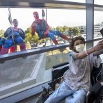 A woman in a hospital gown, cap and mask takes a selfie as three people in superhero costumes pose behind her, on the other side of a window.