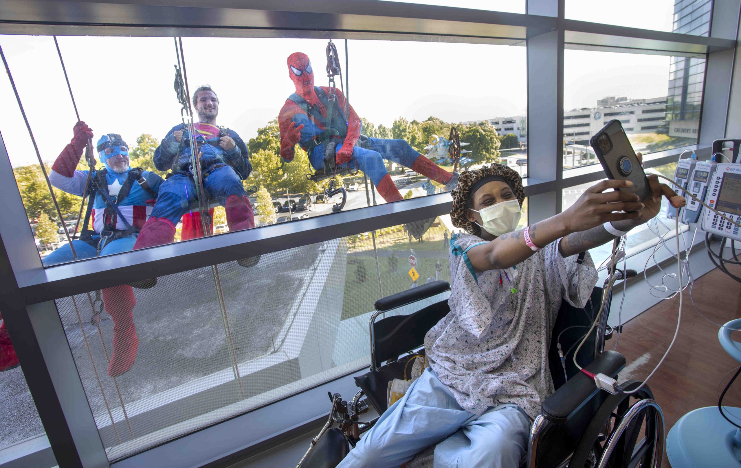 A woman in a hospital gown, cap and mask takes a selfie as three people in superhero costumes pose behind her, on the other side of a window.
