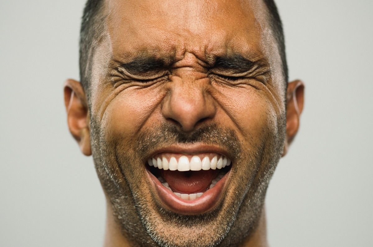 Close up portrait of adult middle eastern man with excited expression and eyes closed against white gray background. Vertical shot of real people from Pakistan laughing with exhilaration in studio with short black hair. Photography from a DSLR camera. Sharp focus on eyes.