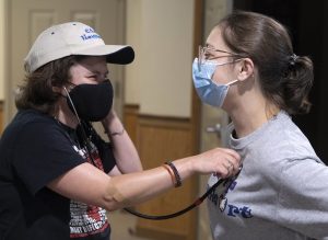 Two young women in masks face one another. One listens to another's heart with a stethoscope.