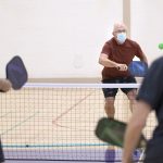 A man in a surgical mask charges a net brandishing a pickleball racquet in a gym. Two other men are seen from behind in the foreground.