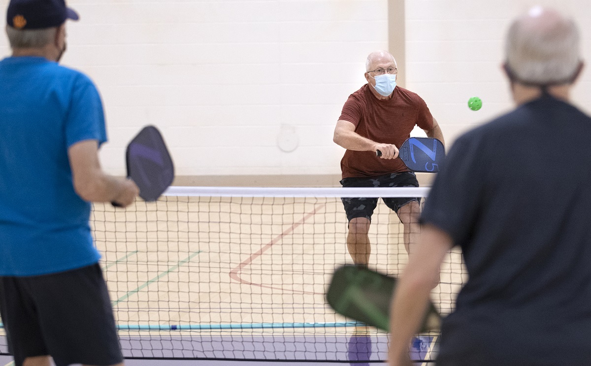A man in a surgical mask charges a net brandishing a pickleball racquet in a gym. Two other men are seen from behind in the foreground.