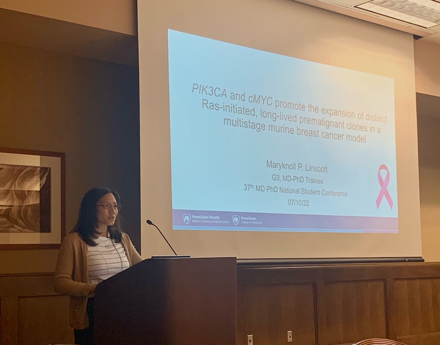 Maryknoll Linscott, MD/PhD student, stands behind a lectern with a microphone, and a large screen to her left shows a slide form her oral presentation.