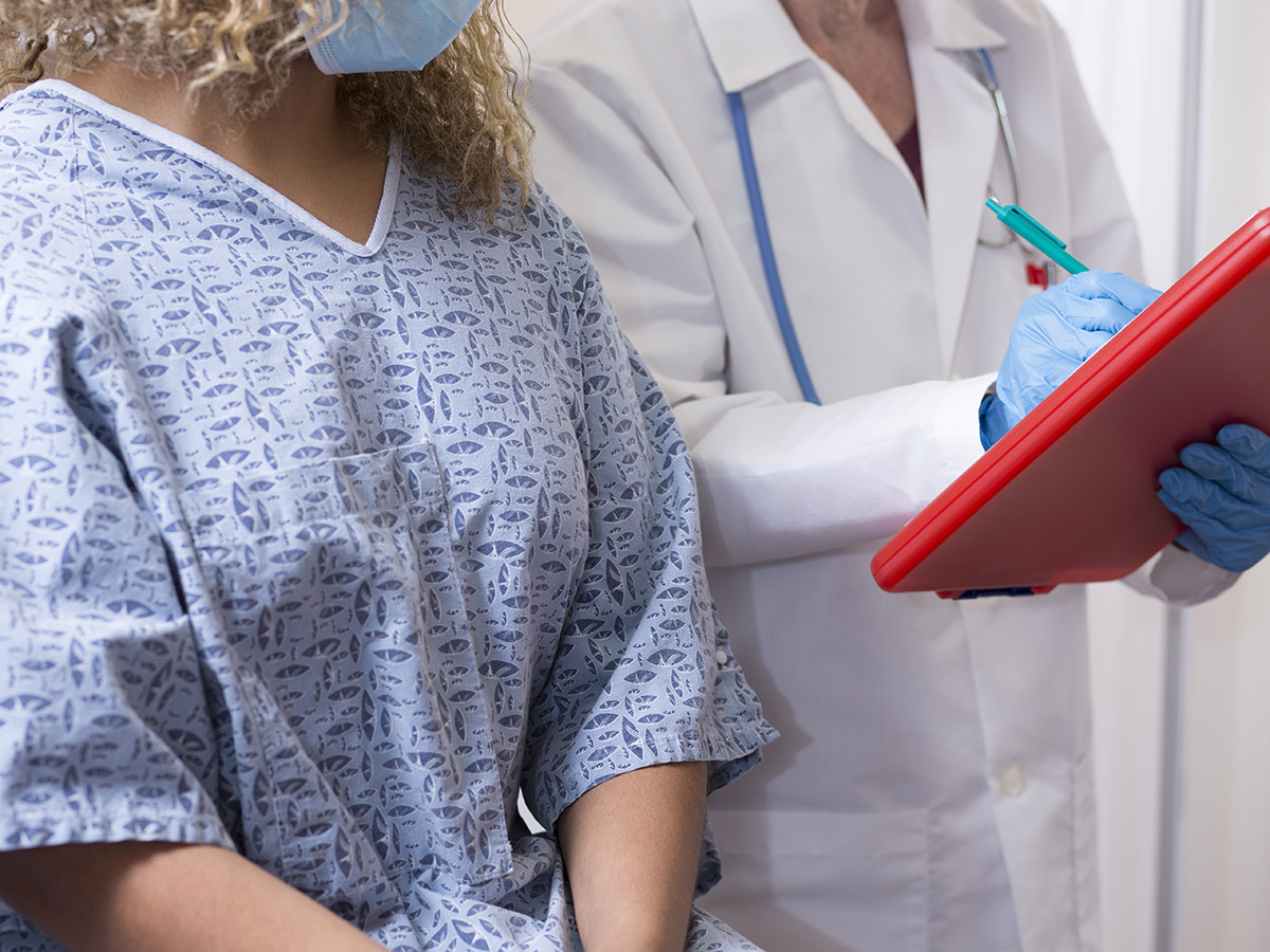 Photo of a doctor and a female patient in an exam room. The patient is wearing a hospital gown while sitting on an exam table, and the doctor is referencing the patient’s medical chart.