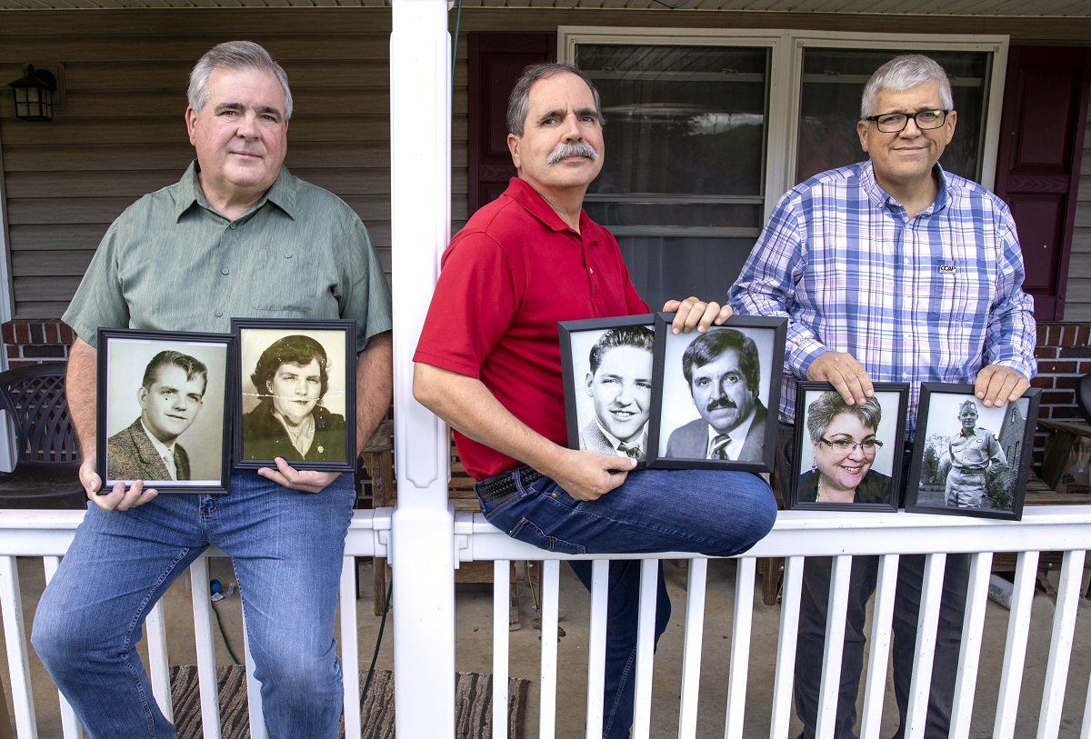 Three men sit and lean on a porch railing, holding framed, black-and-white photos.