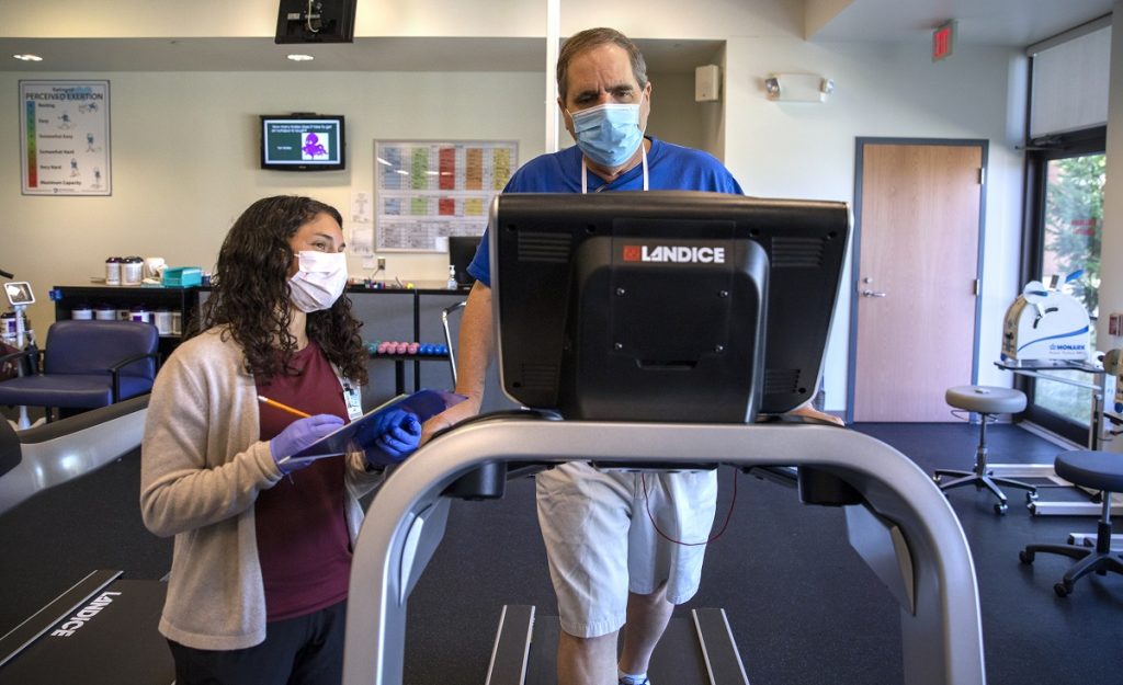 A man in a mask walks on a treadmill. A health care worker in a masks looks at the screen attached to the top of the device.