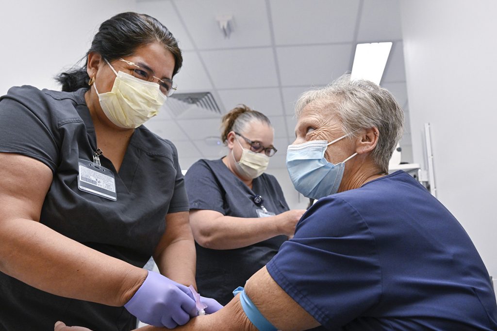 Three women in scrubs and masks are in a room. Two women are standing and one is sitting, while getting her blood drawn.