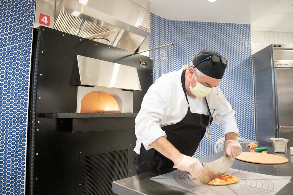 A man cuts a pizza using a large knife. He is wearing a face mask, apron and cap.