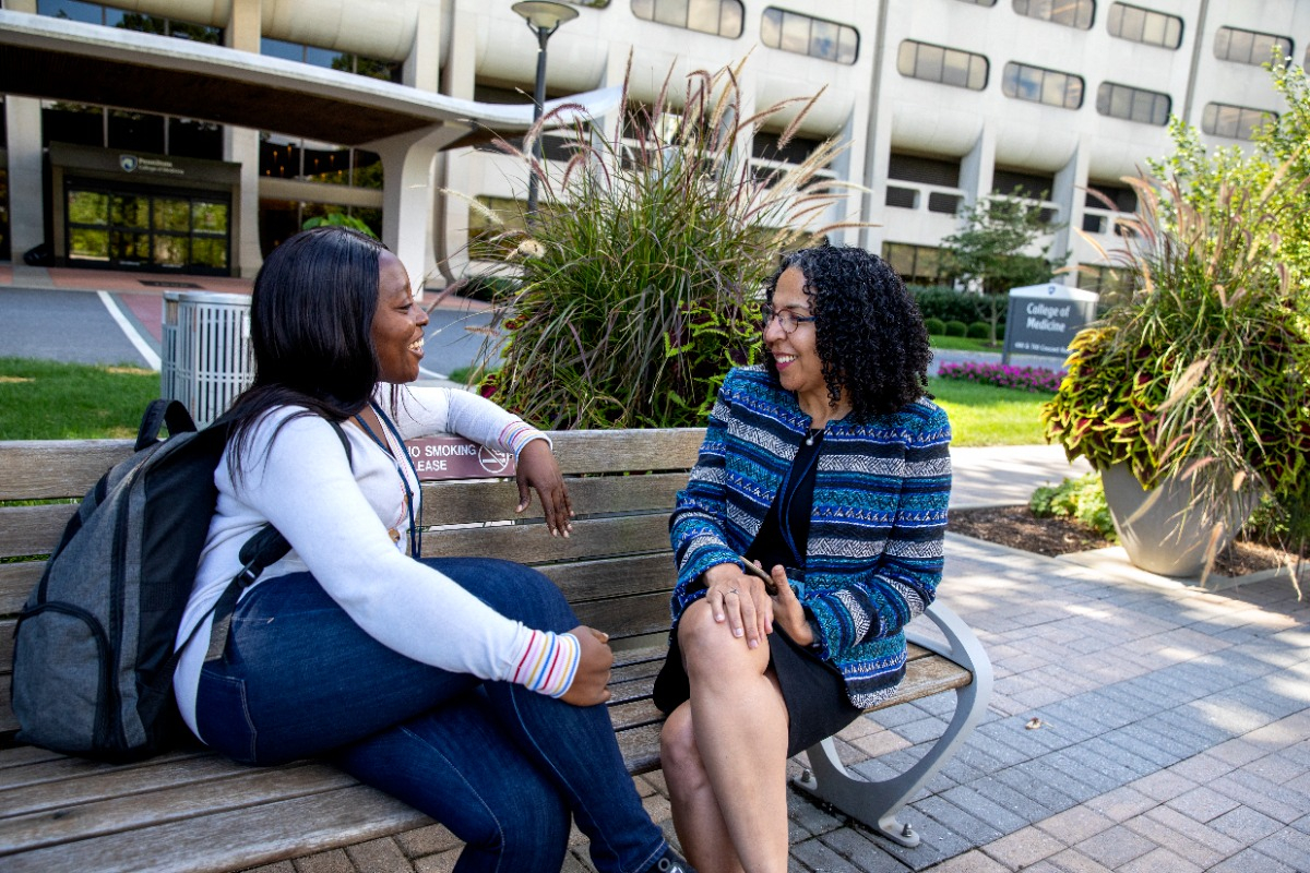 Dr. Inginia Genao sits on a bench outside the Penn State College of Medicine with a student.