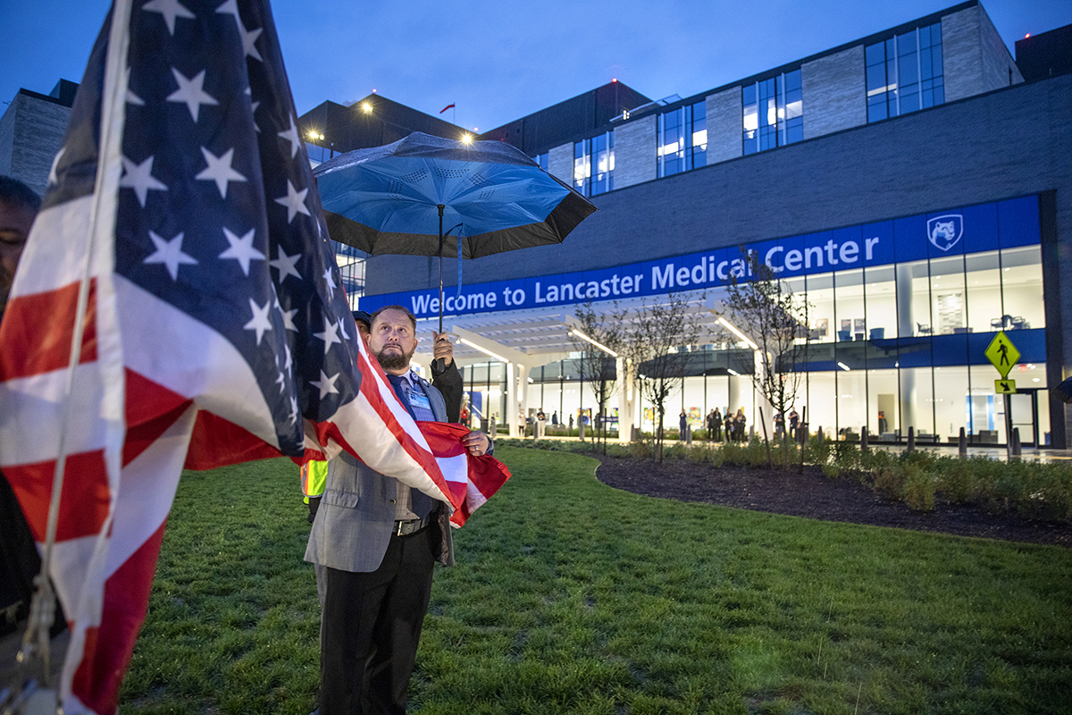 Gentleman under umbrella assists with raising the American flag in front of Lancaster Medical Center building.