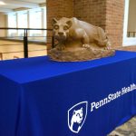 A statue of a lion sits on top of a table. The table is covered with a blue cloth that says, "Penn State Health."