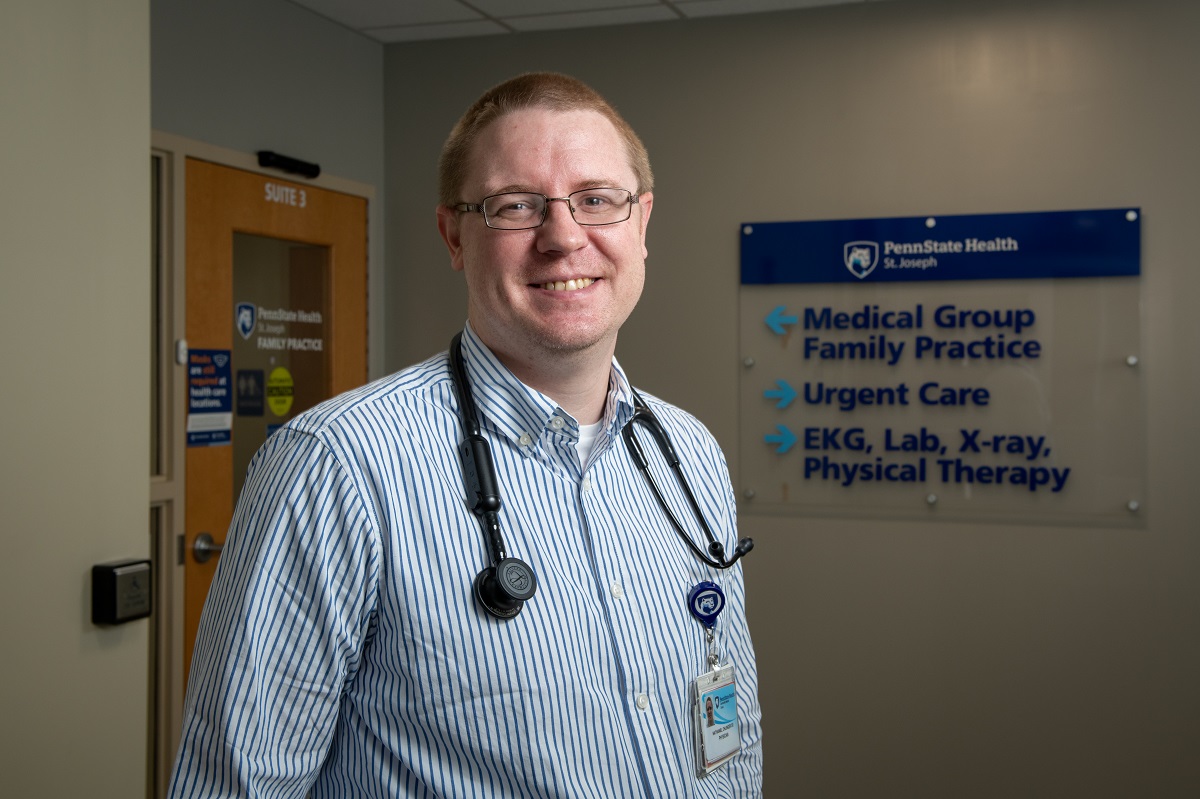 A man with a stethoscope around his neck smiles in a hallway labeled for St. Joseph Medical Center.