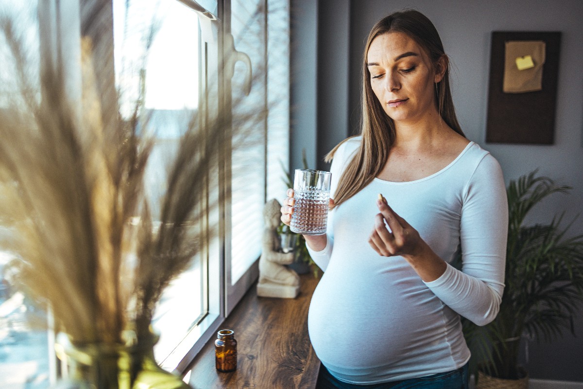 A pregnant woman stands by a window holding a pill in her left hand and a drink in her right hand.