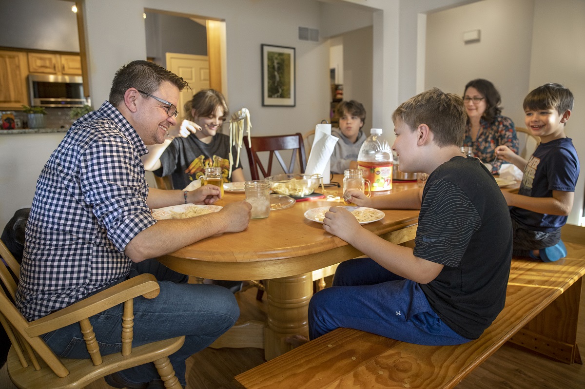 A family enjoys pasta around a dining room table. In the foreground, a father and son laugh together.