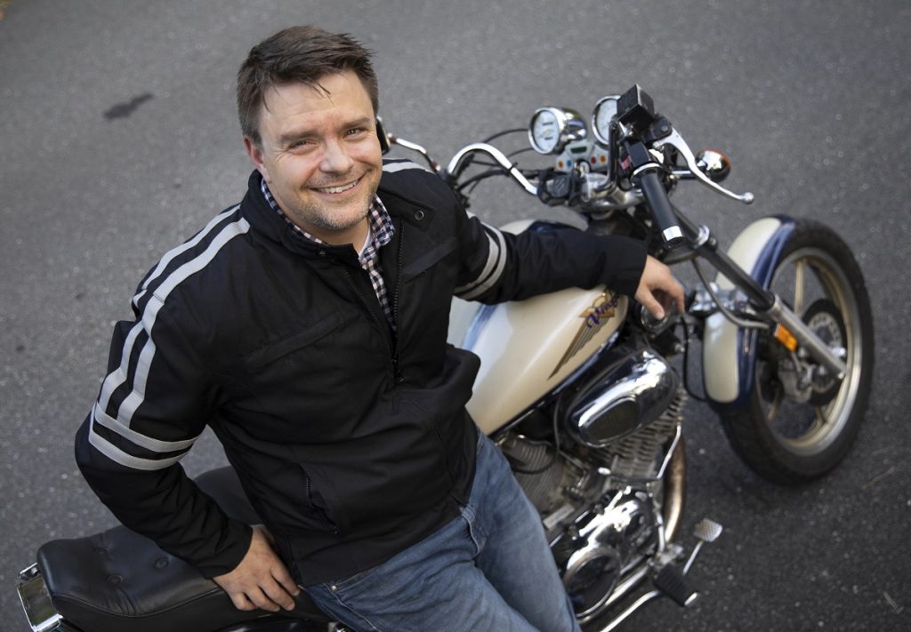 A man smiles at the camera as he leans against a motorcycle.