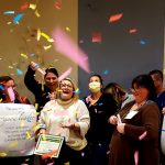 Ticker tape falls from the ceiling as a woman smiles and holds a plaque. She is surrounded by other health care workers, some wearing masks.