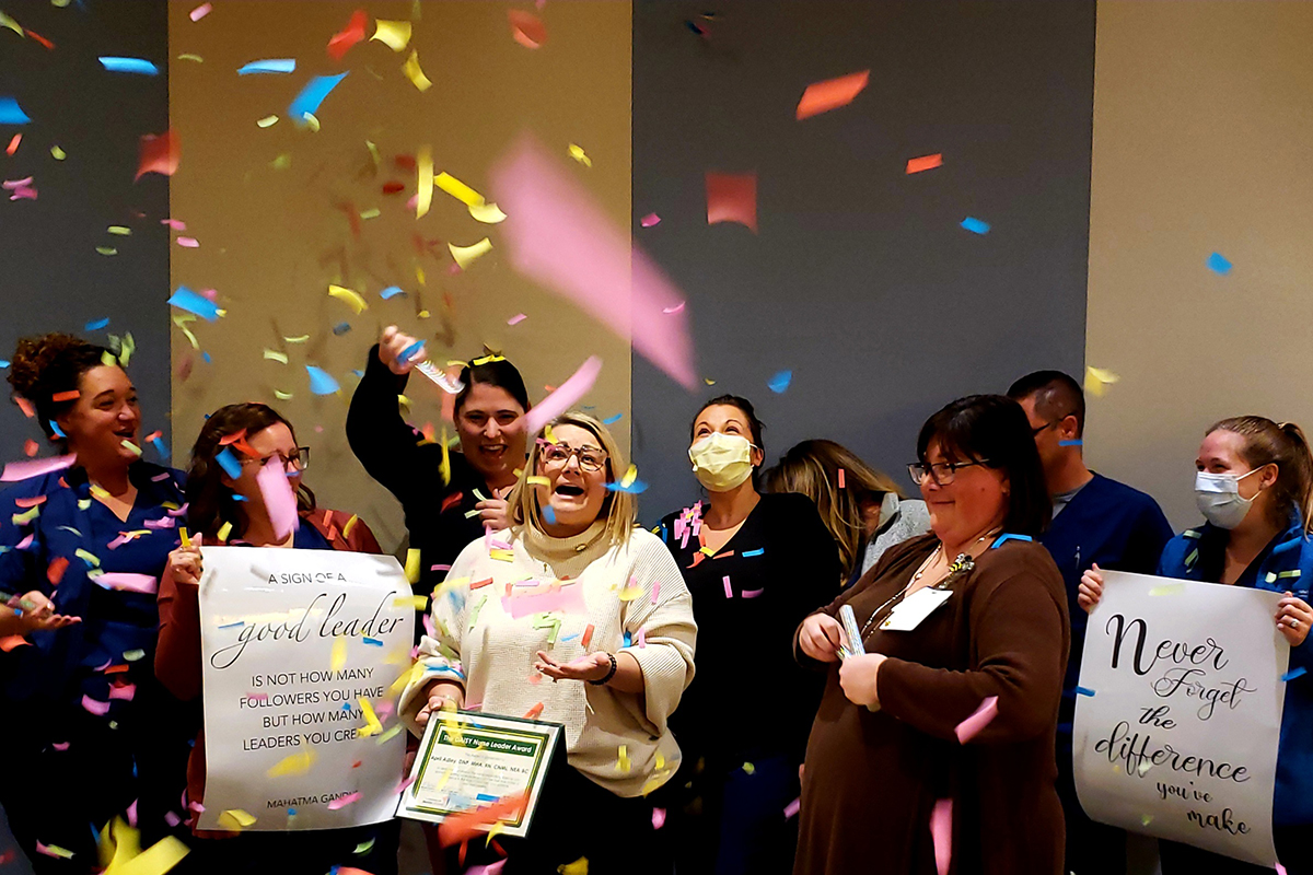 Ticker tape falls from the ceiling as a woman smiles and holds a plaque. She is surrounded by other health care workers, some wearing masks.