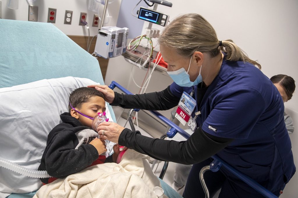Joylyn Meadows, a registered nurse, leans over 5-year-old Rashawn Collazo and adjusts his oxygen mask in the Emergency Department of Penn State Health Lancaster Medical Center. She is wearing scrubs, a name badge and a face mask. Collazo is propped up in bed and is wearing a hooded sweatshirt. 