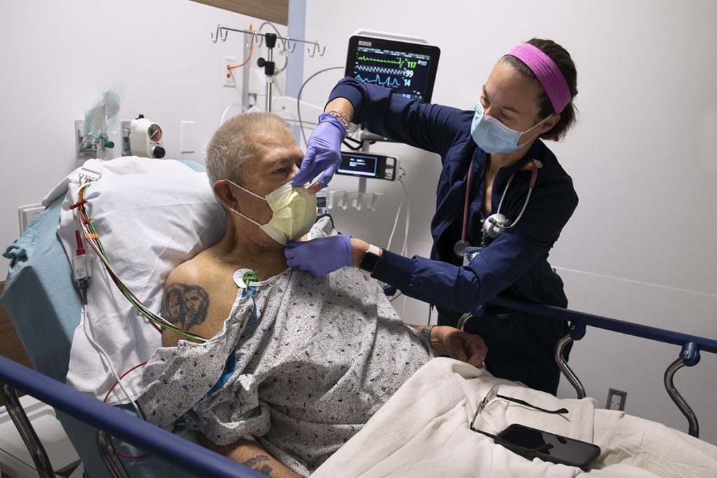 Brittney Patterson, a registered nurse, adjusts the mask of Daniel Rutledge during his visit to the Emergency Department at Penn State Health Lancaster Medical Center. Patterson is wearing scrubs, a face mask and has a stethoscope around her neck. Rutledge is sitting up in bed and wearing a hospital gown. 