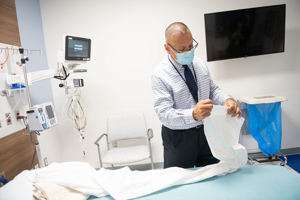Esteban Monserrat, Sodexo senior supervisor for environmental health services, unfolds a sheet over a bed in the Emergency Department at Penn State Health Lancaster Medical Center. He is wearing a shirt and tie, face mask and glasses. 