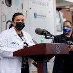 Dr. Sarah Ramirez of Penn State Health stands at a podium next to a man and woman during a press conference at Beacon Clinic in Harrisburg. She is wearing a white coat and a face mask. Behind her is a van with the Highmark logo on it.