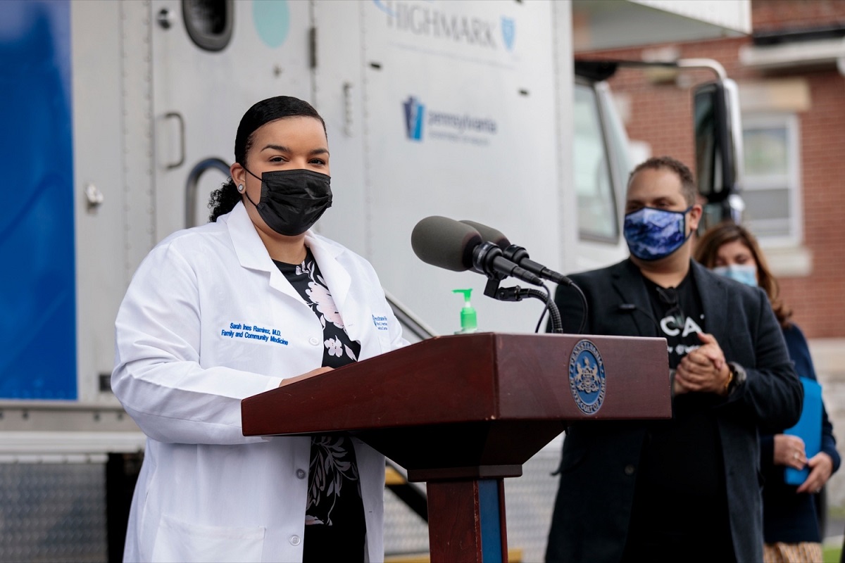 Dr. Sarah Ramirez of Penn State Health stands at a podium next to a man and woman during a press conference at Beacon Clinic in Harrisburg. She is wearing a white coat and a face mask. Behind her is a van with the Highmark logo on it.