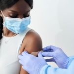 Image features a woman in a mask receiving a bandage after an injection.