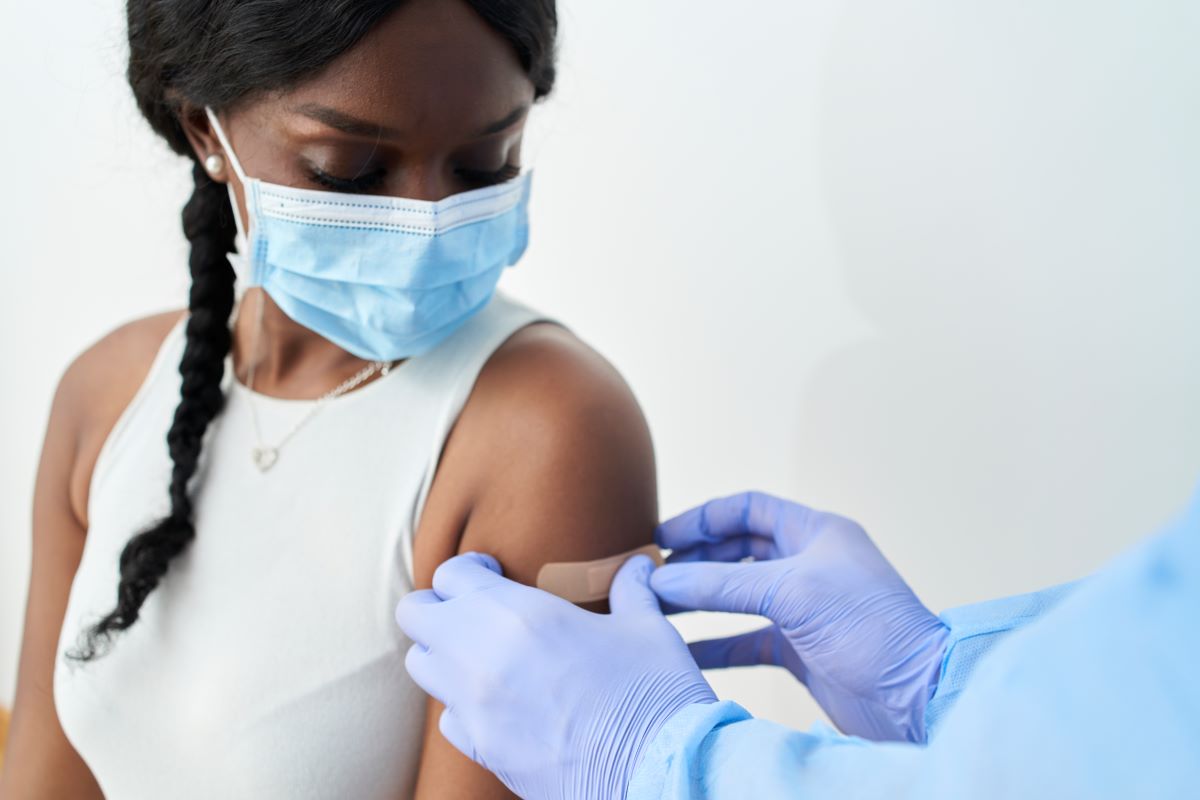 Image features a woman in a mask receiving a bandage after an injection.