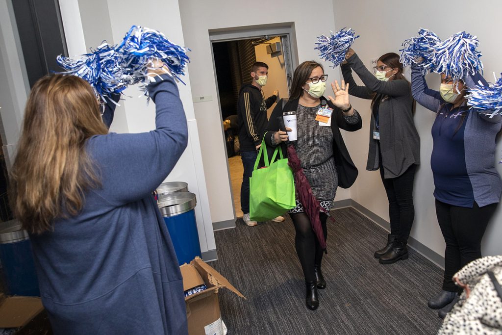 A woman walks through a doorway while coworkers cheer her on with pom-poms.