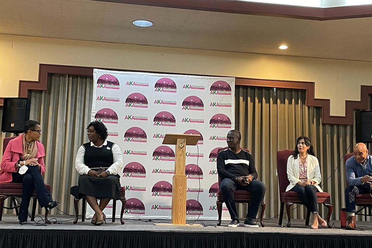 A row of five people sit on a stage. At the center is a podium and behind them a logo appears on a poster.