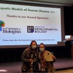 Two masked women stand together. One holds an award.