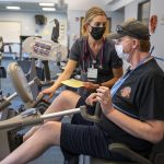 A man in a ball cap and shorts works on a piece of equipment in a gym. A woman in a surgical mask stands next to him.
