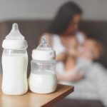 Two bottles of breast milk sit on a counter. Faded into the background, a mother breastfeeds her baby.