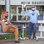 Eddie Holman stands in front of Penn State Health Milton S. Hershey Medical Center with an IV pole and monitor. He is wearing a Penn State T-shirt and pants, a surgical mask and glasses. Next to him is a statue of the Penn State Nittany Lion sitting on a bench.