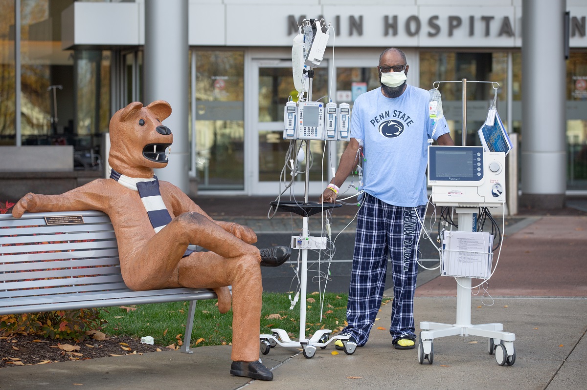 Eddie Holman stands in front of Penn State Health Milton S. Hershey Medical Center with an IV pole and monitor. He is wearing a Penn State T-shirt and pants, a surgical mask and glasses. Next to him is a statue of the Penn State Nittany Lion sitting on a bench.