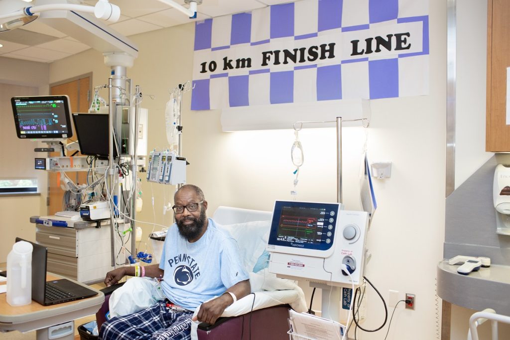 Eddie Holman sits in a chair in his room at the Milton S. Hershey Medical Center. He is hooked up to an IV and a heart monitor is on his right. He is wearing a Penn State T-shirt, pants and glasses. Behind him is a sign that reads “10 km finish line.” 
