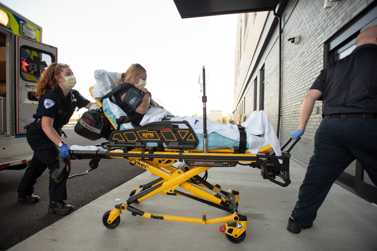 A woman sits upright on a litter as two ambulance staff members guide it from the ambulance to the sliding door entryway of a hospital.