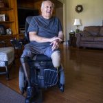 A man sits in a wheelchair a living room. His right leg is a prosthetic. His left is amputated near the knee. The man smiles and speaks.