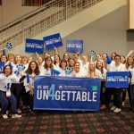 A group of people in white shirts waving signs that say "Un4gettable" stand in a carpeted room in front of a stair case.